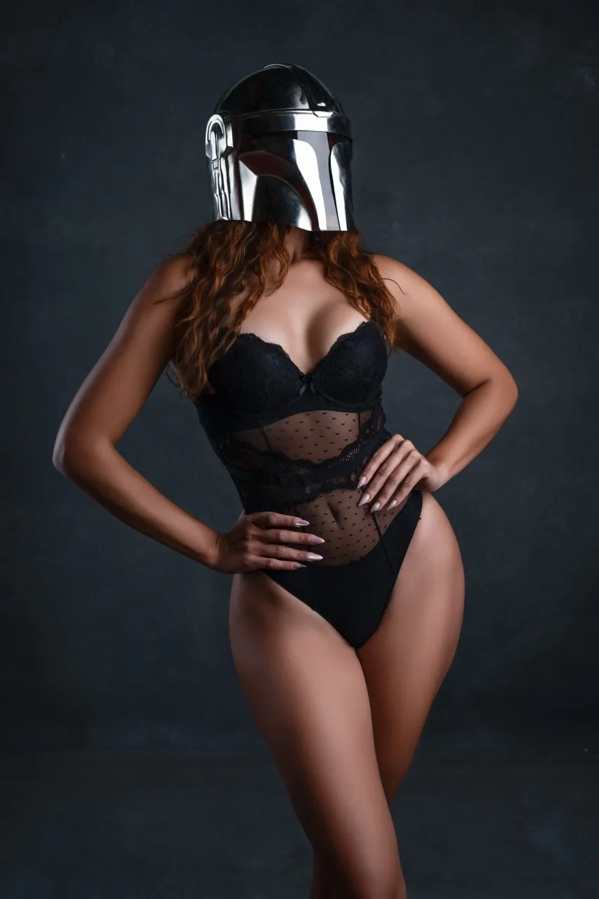 Boudoir model wearing black lace lingerie and a Mandalorian helmet, striking a confident pose against a dark textured backdrop—blending sci-fi edge with sensual elegance.