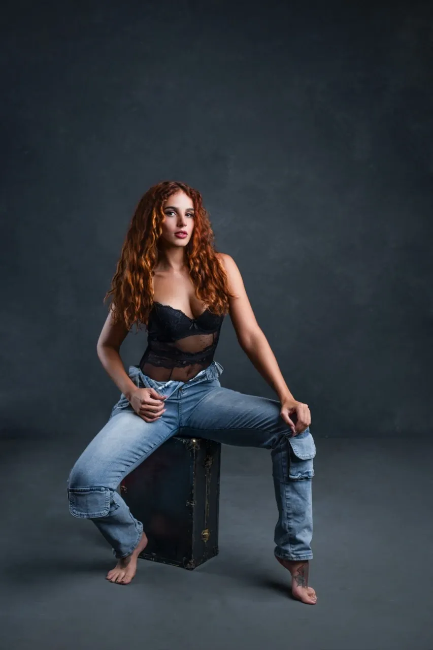 Boudoir model wearing jeans and black lace lingerie, seated confidently with a slight waistband pull—suggestive and stylish against a textured gray backdrop.