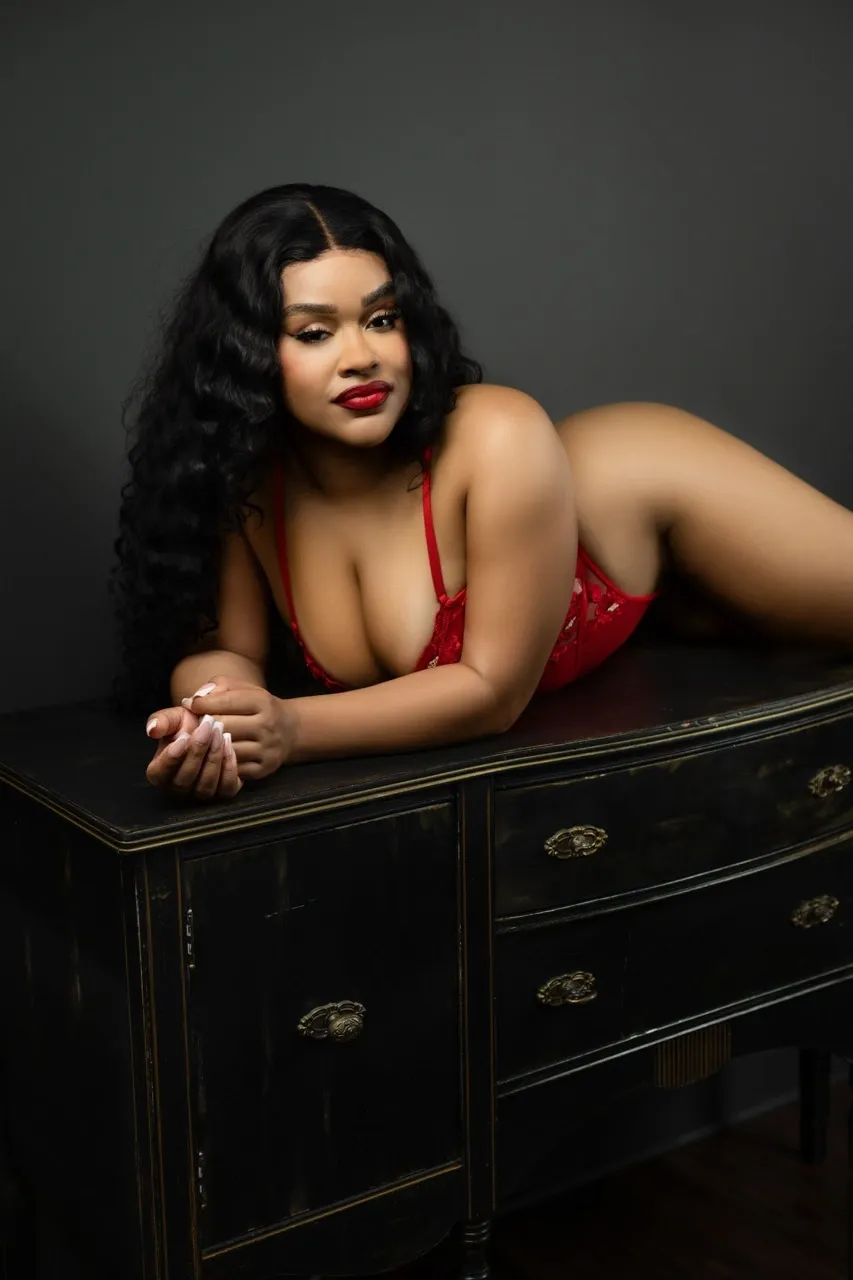 Confident Black woman in red lace lingerie posing on vintage black vanity in moody boudoir studio with dark gray backdrop.