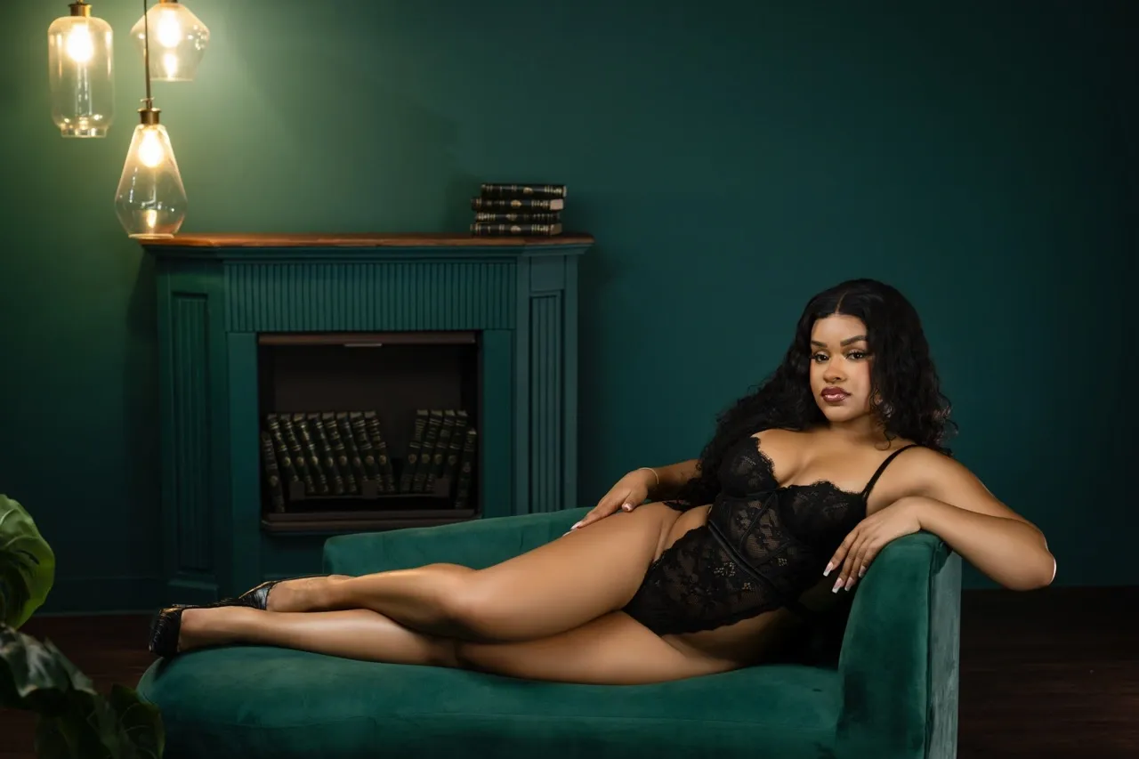 Stunning Black woman in black lace bodysuit lounging confidently on emerald green velvet couch in a moody boudoir studio with vintage lighting.
